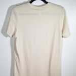 the back of a cream t-shirt on hanger hanging on a nail against a white wall.