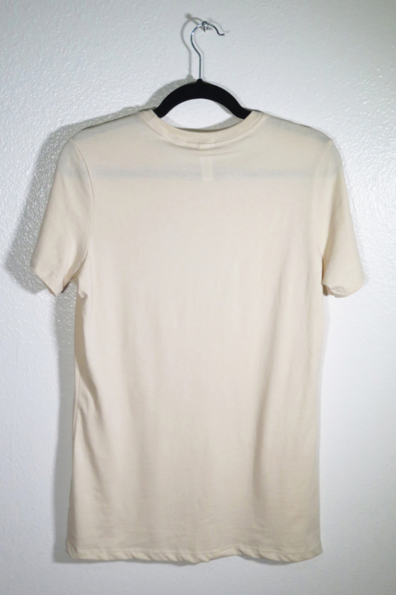 the back of a cream t-shirt on hanger hanging on a nail against a white wall.