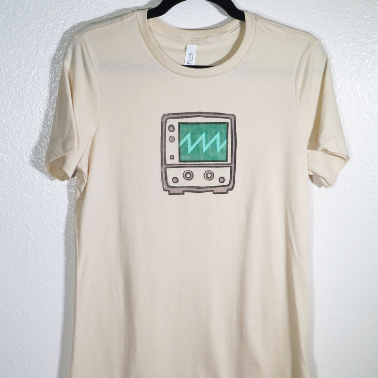 cream t-shirt on hanger hanging on a nail against a white wall. A block print of an oscilloscope displaying a sawtooth waveform is hand printed centered over the chest area.