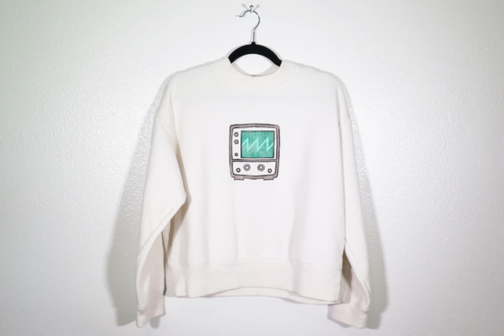 off-white sweatshirt on hanger hanging on a nail against a white wall. A block print of an oscilloscope displaying a sawtooth waveform is hand printed centered over the front chest area.