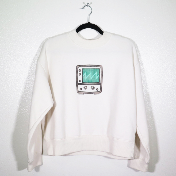 off-white sweatshirt on hanger hanging on a nail against a white wall. A block print of an oscilloscope displaying a sawtooth waveform is hand printed centered over the front chest area.