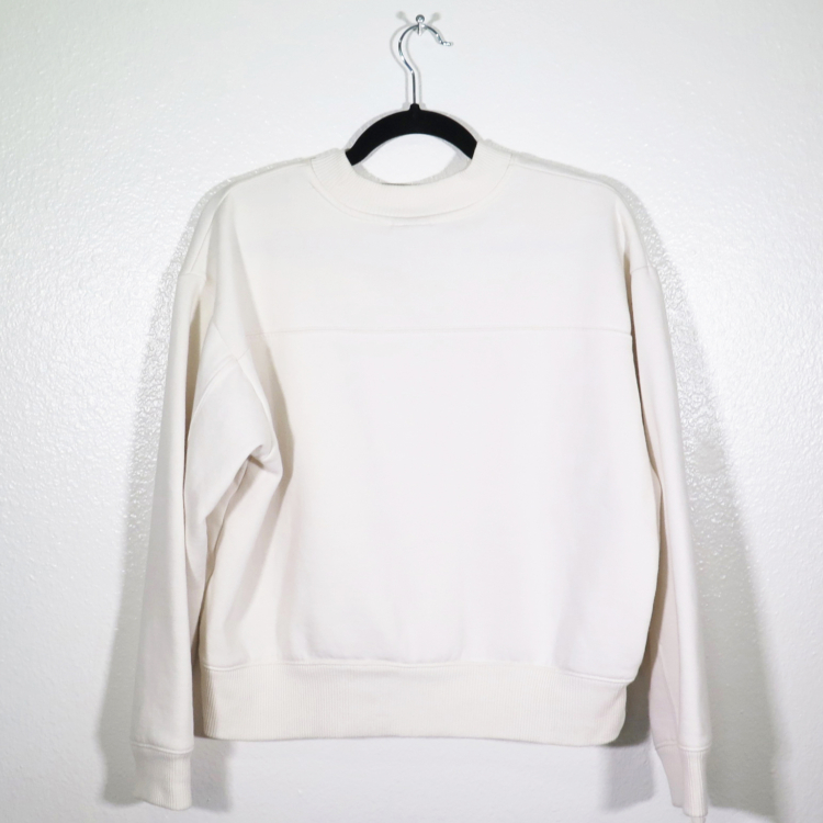 off-white sweatshirt on hanger hanging on a nail against a white wall with the back facing the viewer.