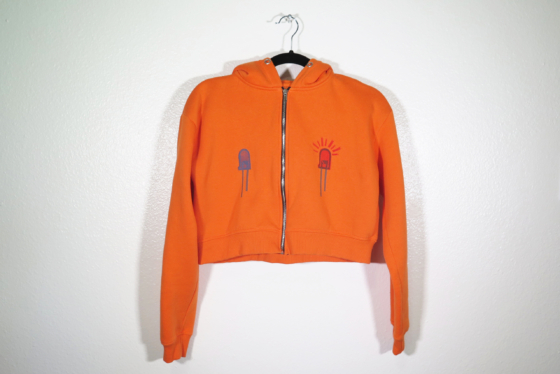 bright orange sweatshirt on hanger hanging on a nail against a white wall. Two LEDs are hand printed onto front chest area, one blue and one red.