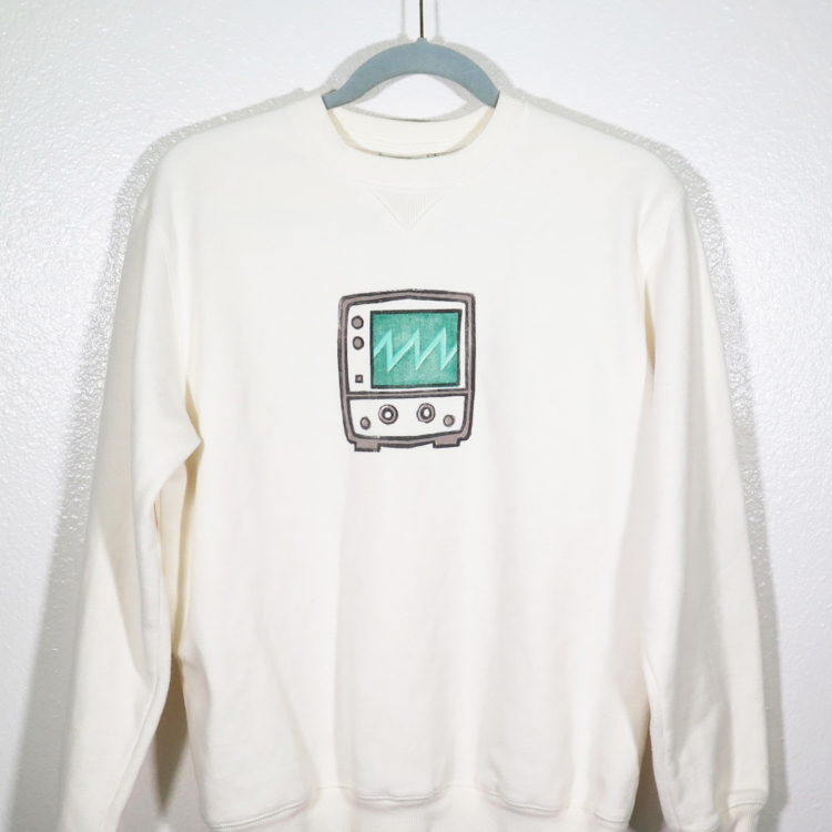 cream sweatshirt on hanger hanging on a nail against a white wall. A block print of an oscilloscope displaying a sawtooth waveform is hand printed centered over the front chest area.