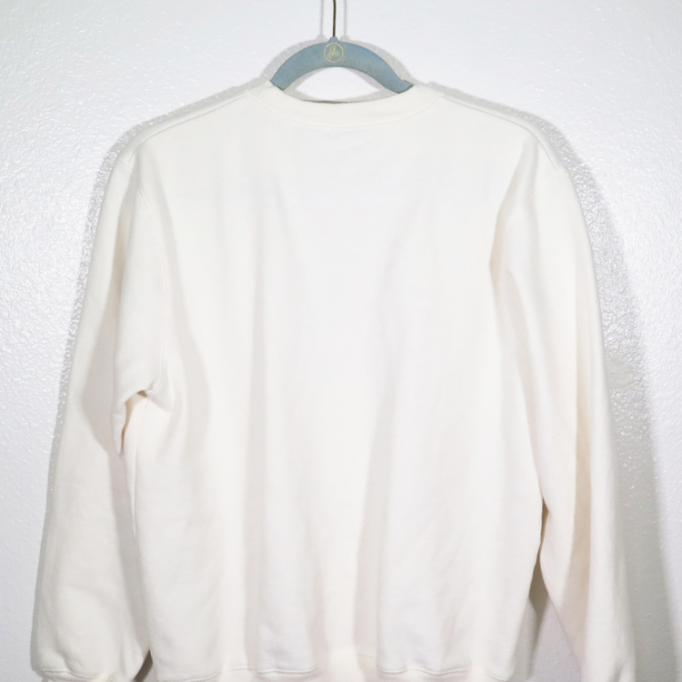 cream sweatshirt on hanger hanging on a nail against a white wall with the back facing the viewer.