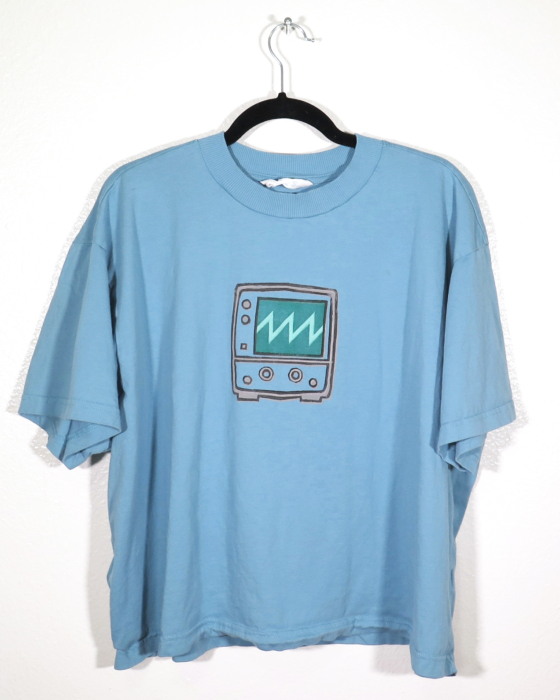 blue t-shirt on hanger hanging on a nail against a white wall. A block print of an oscilloscope displaying a sawtooth waveform is hand printed centered over the chest area.