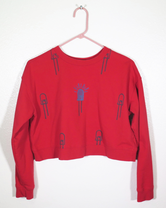 front of red sweatshirt on hanger hanging on a nail against a white wall. It is hand printed with an LED design