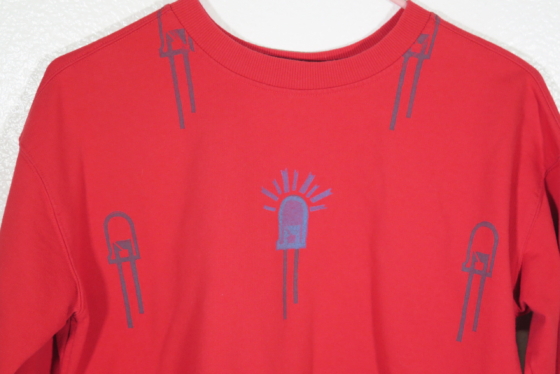 close up of red sweatshirt on hanger hanging on a nail against a white wall. It is hand printed with an LED design