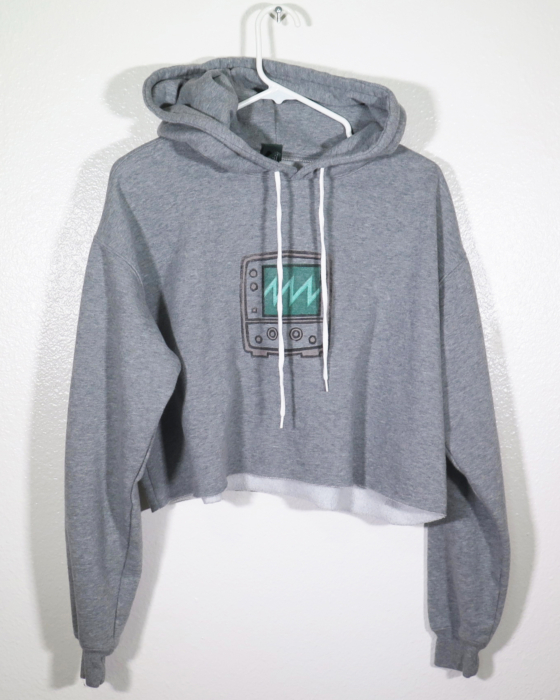 The front of a gray hooded sweatshirt that has long sleeves and is cropped on a hanger. The hanger is hanging off a nail against a while wall. A block print of an oscilloscope showing a sawtooth wave is hand printed in the center front.