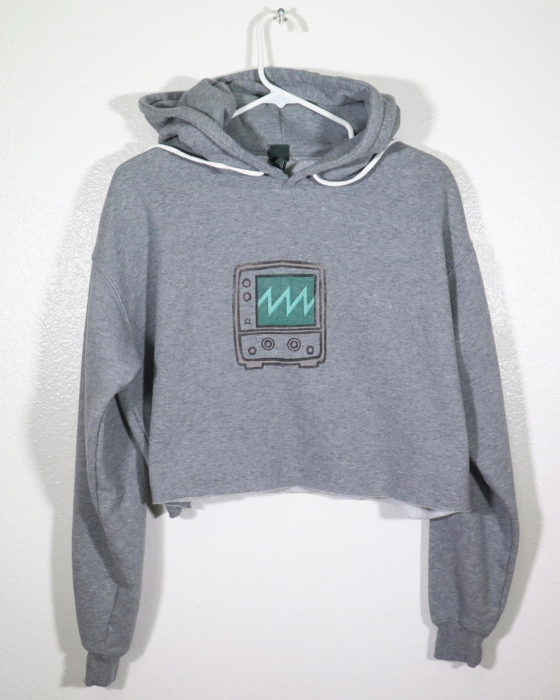 Alternative view of the front of a gray hooded sweatshirt with long sleeves and is cropped on a hanger. The hanger is hanging off a nail against a while wall. A block print of an oscilloscope showing a sawtooth wave is hand printed in the center front. This version has the hoodie strings up to better show the print.