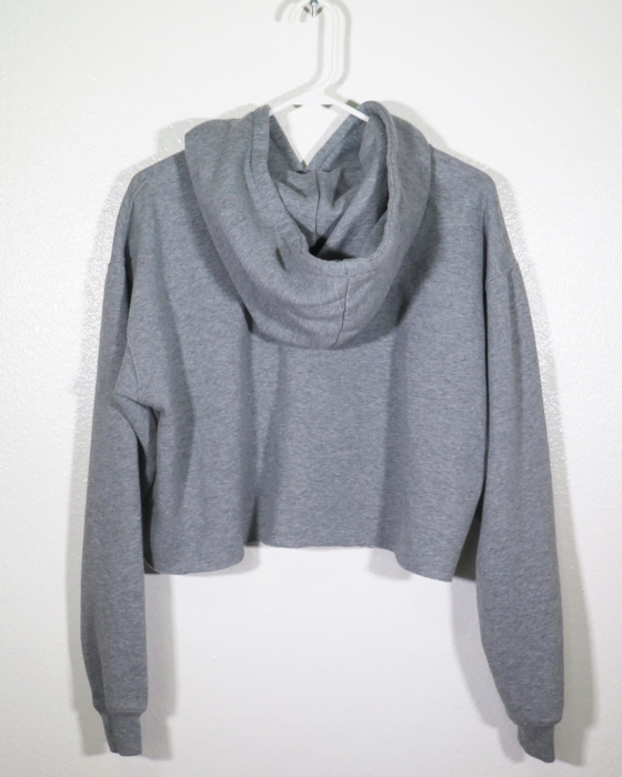 Back of a gray hooded sweatshirt with long sleeves and is cropped. It is on a hanger hanging off a nail against a while wall.
