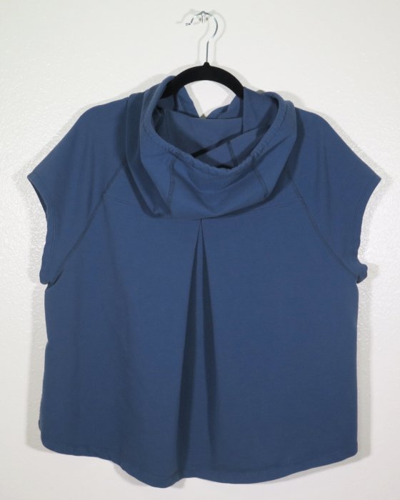 A navy sleeveless hooded shirt on a hanger hangs on a nail against a white wall. Showing the back that has a pleat down the center back.