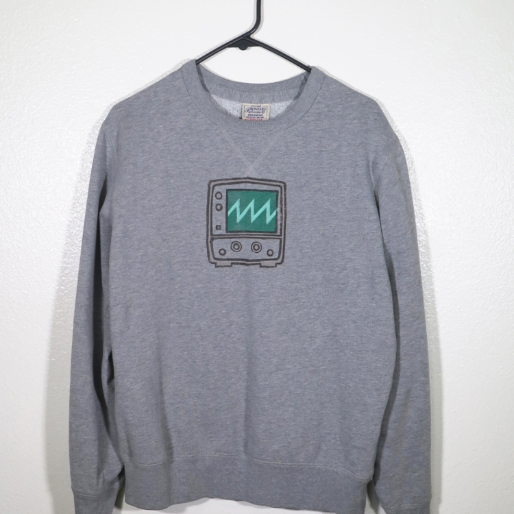 Front view of gray long sleeved sweatshirt on a hanger hanging on a nail in front of a white wall. A block print of an oscilloscope displaying a sawtooth wave is hand printed on over the chest area.