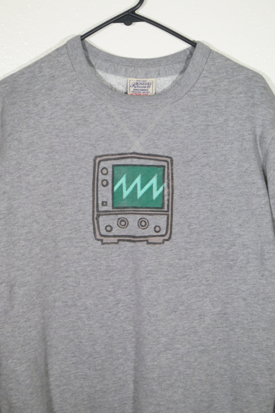 Medium close view of the front of gray long sleeved sweatshirt on a hanger hanging on a nail in front of a white wall. A block print of an oscilloscope displaying a sawtooth wave is hand printed on over the chest area.