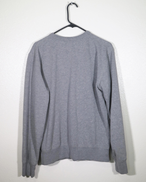 Back view of gray long sleeved sweatshirt on a hanger hanging on a nail in front of a white wall.
