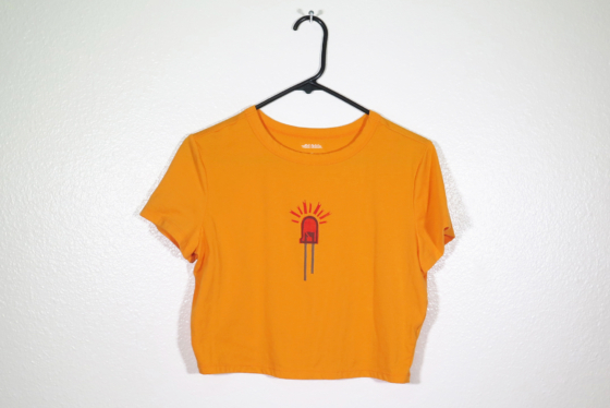 front of orange t-shirt on hanger hanging on a nail against a white wall. It is hand printed with an LED design