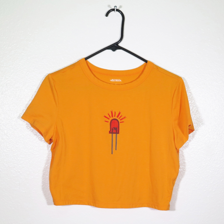 front of orange t-shirt on hanger hanging on a nail against a white wall. It is hand printed with an LED design