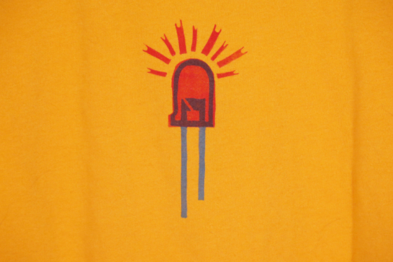 close up of the front of an orange t-shirt on hanger hanging on a nail against a white wall. It is hand printed with an LED design