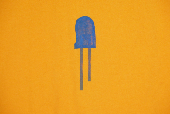 close up of the back of an orange t-shirt on hanger hanging on a nail against a white wall. It is hand printed with an LED design