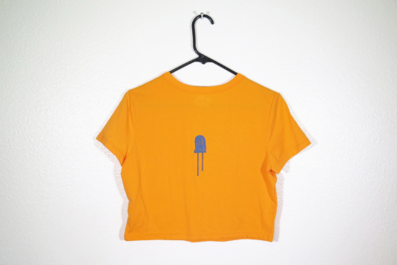 back of orange t-shirt on hanger hanging on a nail against a white wall. It is hand printed with an LED design