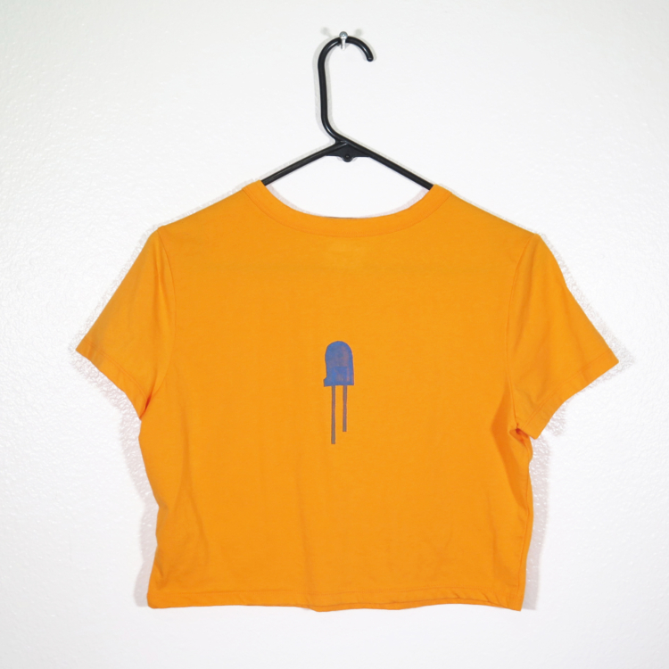 back of orange t-shirt on hanger hanging on a nail against a white wall. It is hand printed with an LED design