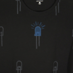 close up of black t-shirt on hanger hanging on a nail against a white wall. It is hand printed with an LED design