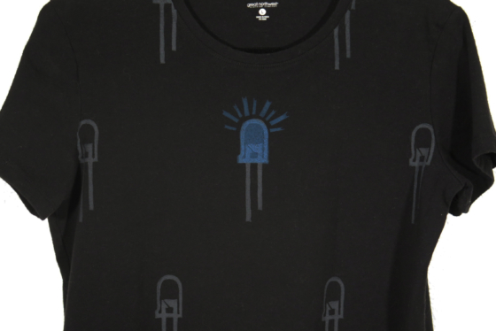close up of black t-shirt on hanger hanging on a nail against a white wall. It is hand printed with an LED design