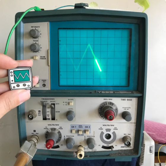 A 1976 Tektronix oscilloscope with a triangle wave being displayed on its old CRT screen. A hand holds up an enamel pin design of the oscilloscope, connecting the design to the inspiration.